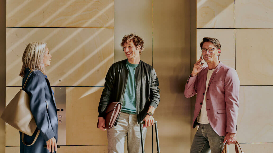 A businesswoman and two businessmen, one with a smartphone to his ear, wait in front of an elevator