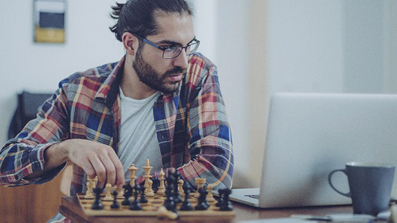 Ein junger Mann sitzt zuhause vor seinem Laptop, daneben steht ein Schachbrett. Er lernt Online-Schach.